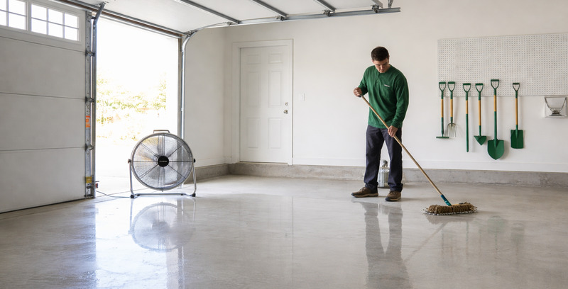 Person mopping a wet concrete garage floor with tools on the wall and a large fan nearby — concrete cleaning.